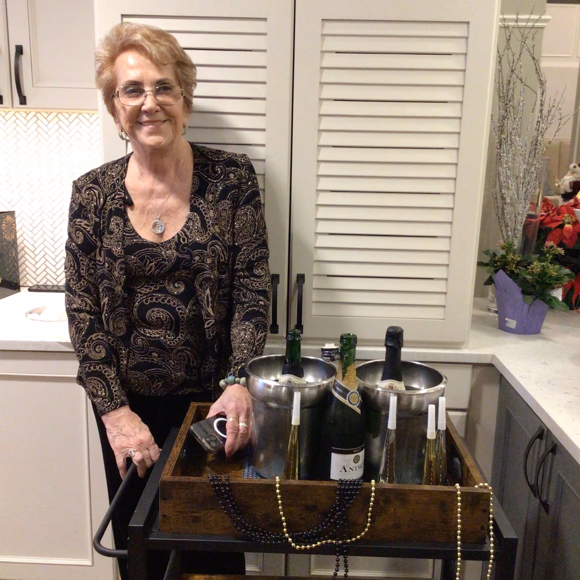 An elderly woman with short blonde hair and glasses stands behind a serving cart in a kitchen area. The cart holds two metal ice buckets with bottles of champagne and other beverages, along with some decorative beads draped over the front. The background shows white cabinets, a countertop with a vase of flowers, and a tiled backsplash.