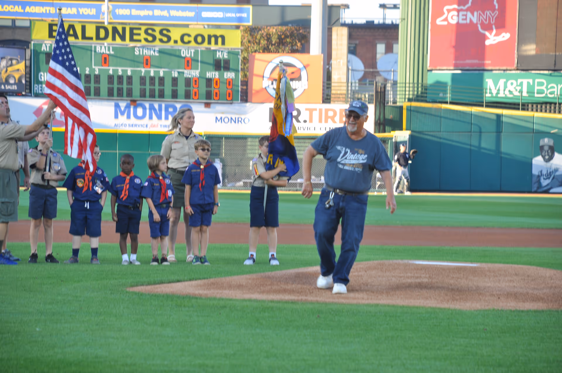 A group of children in scout uniforms standing on a baseball field near the pitcher's mound. One adult is holding an American flag, and another adult is walking on the mound wearing a blue shirt and cap. The scoreboard and advertisements are visible in the background.