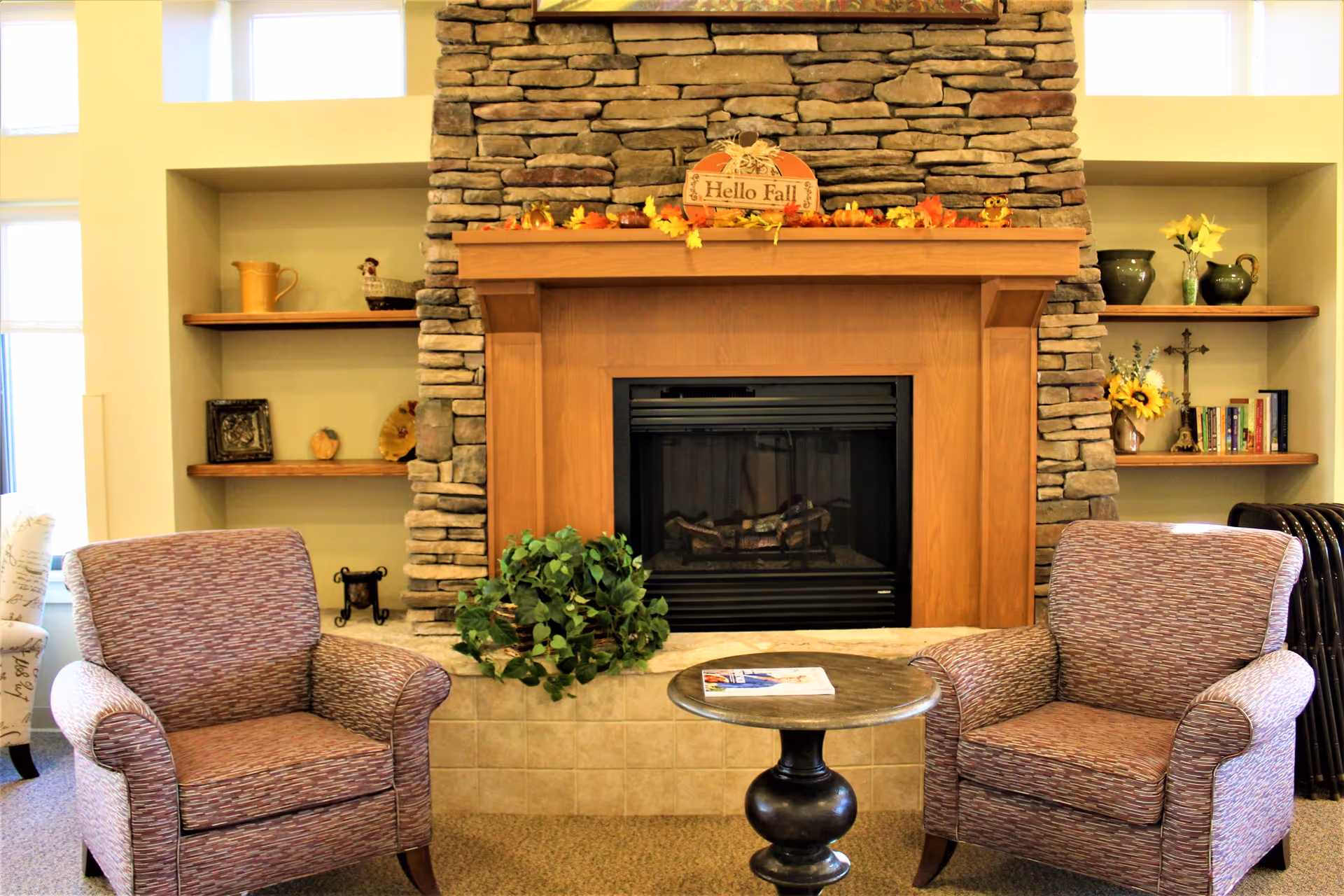 Cozy seating area with two upholstered armchairs facing a stone-surrounded fireplace topped by a wooden mantel and a small round table between them.