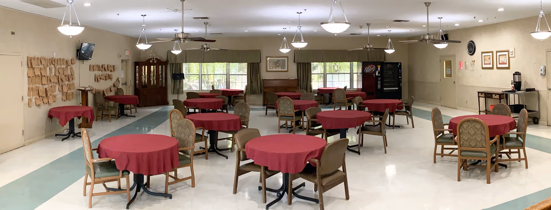 A spacious dining room with multiple round tables covered with red tablecloths, each surrounded by four chairs. The room has large windows with green curtains, ceiling fans, and hanging light fixtures. There is a vending machine and a coffee station along the walls, and a bulletin board with papers pinned on it.