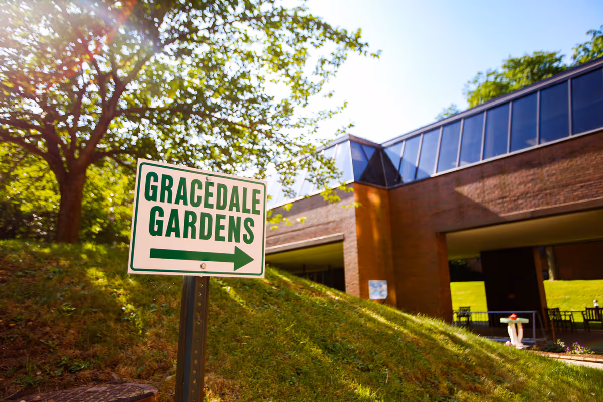 A sign reading "GRACEDALE GARDENS" with an arrow in front of a grassy slope and a brick building under trees.