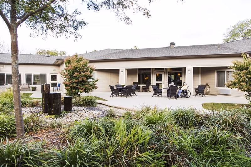 Front patio and entrance of an assisted living building with outdoor seating, landscaping, and a person in a wheelchair.