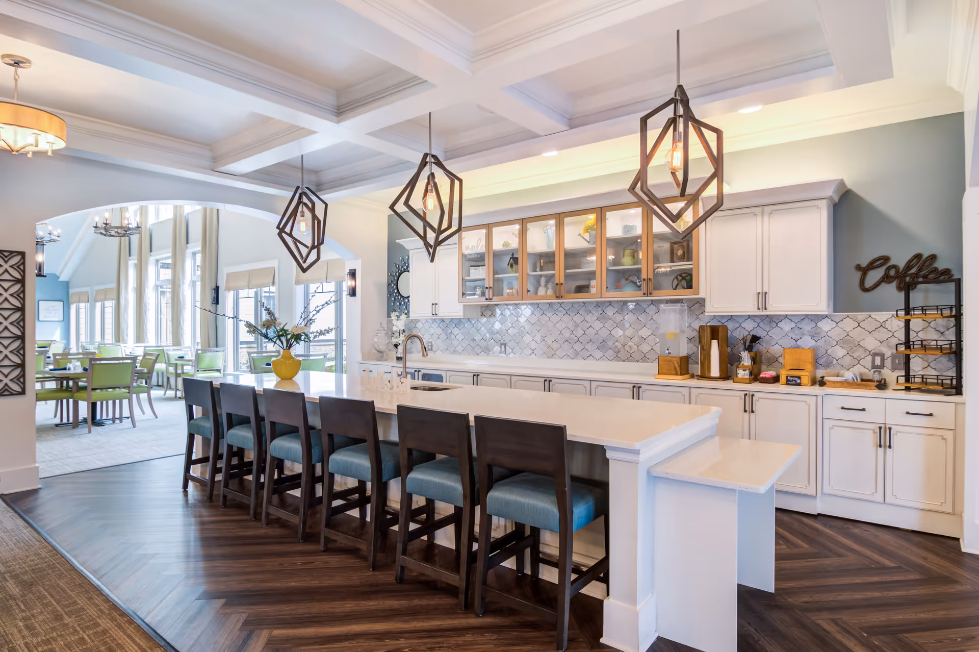 A modern kitchen area with a long white island countertop surrounded by six dark wooden chairs with teal cushions. Above the island hang three geometric pendant lights. The back wall features white cabinets with glass doors on the upper section and a decorative tile backsplash. To the right, there is a coffee station with a sign that says 'Coffee'. In the background, a dining area with green chairs and large windows is visible through an arched opening.