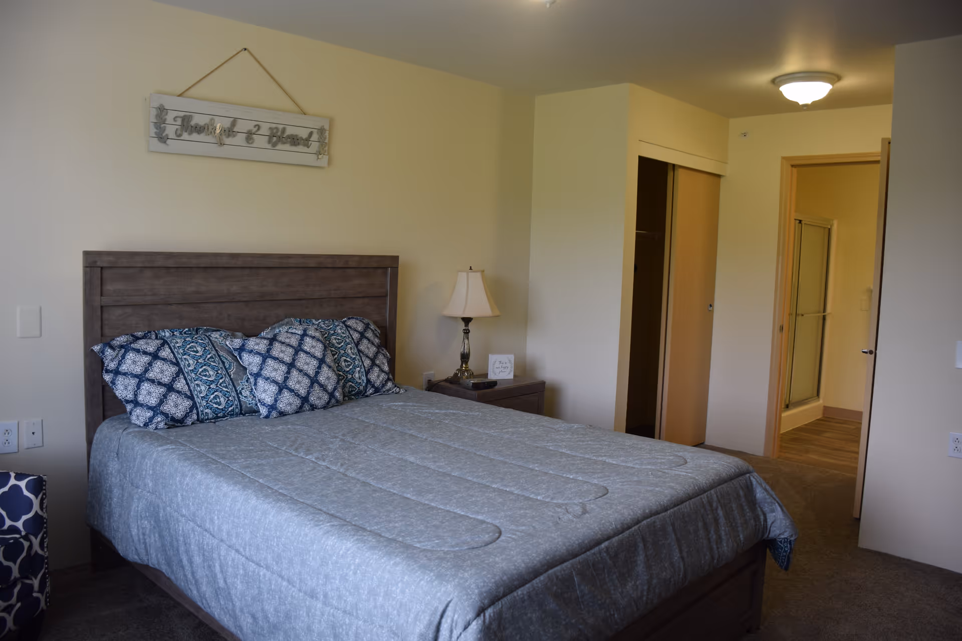 A bedroom with a wooden bed frame and a gray quilted bedspread. There are three decorative pillows with blue and white patterns on the bed. A nightstand with a lamp and a small framed sign is next to the bed. On the wall above the bed is a decorative sign that reads 'Thankful & Blessed'. In the background, there is an open closet with sliding doors and a doorway leading to a bathroom with a glass shower door.