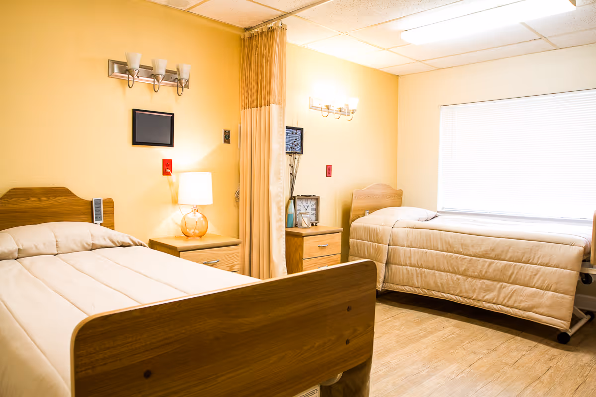 A bright room with two single beds separated by a privacy curtain. Each bed has a wooden headboard and beige bedding. There are two wooden nightstands, one with a lamp and the other with a clock and decorative items. The room has light yellow walls, a window with blinds, and ceiling lights.