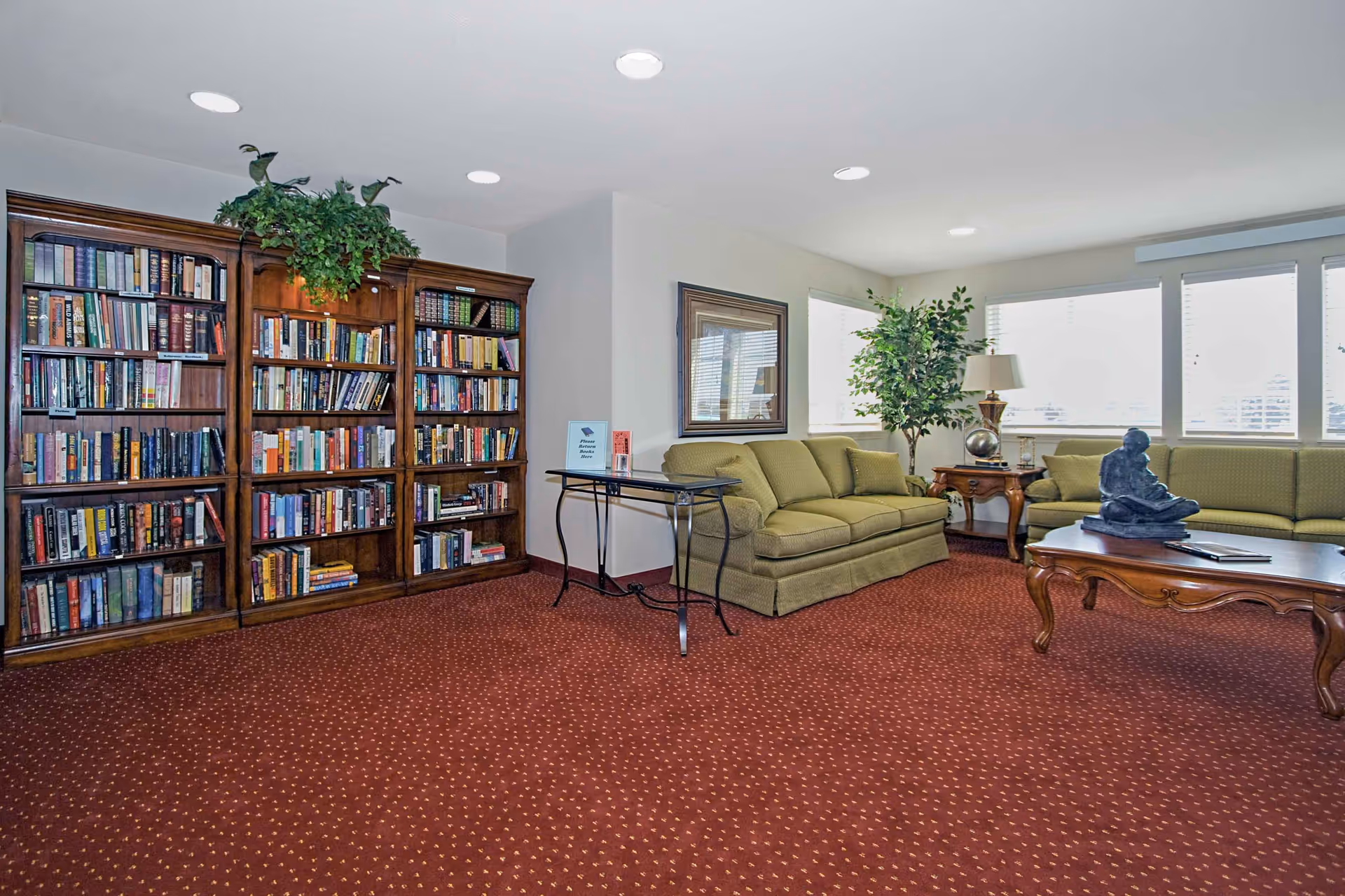 A cozy living room area with two green sofas, a wooden coffee table with a sculpture on it, a wooden side table with a lamp and a plant, a large wooden bookshelf filled with books, and a small table with a sign that says 'Please Return Books Here'. The room has a red carpet with a subtle pattern and large windows with blinds letting in natural light.