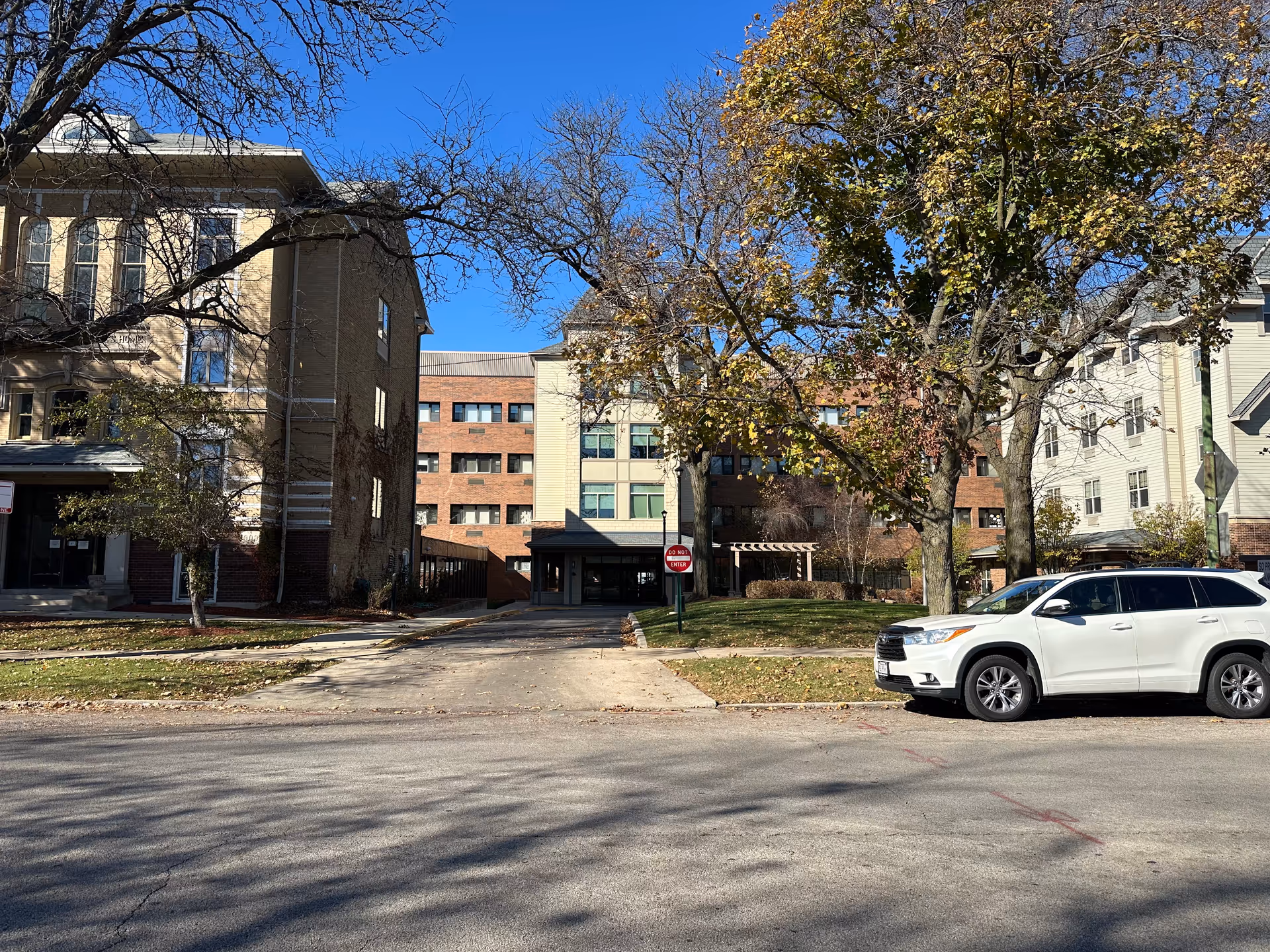 Exterior view of Norwood Crossing facility showing a multi-story building with a driveway entrance, surrounded by trees with autumn foliage and a white SUV parked on the street in front.