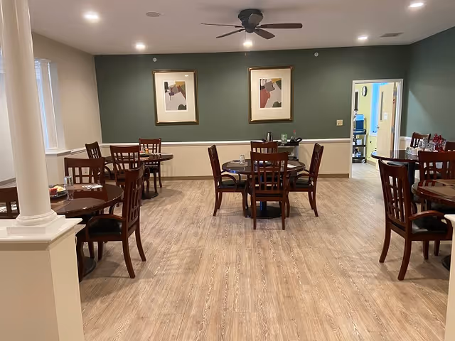 A dining room with several round wooden tables and chairs arranged neatly on a light wood floor. The walls are painted green and beige with two abstract framed artworks hanging on the green wall. A ceiling fan with lights is mounted on the ceiling. There is an open doorway leading to another room in the background.
