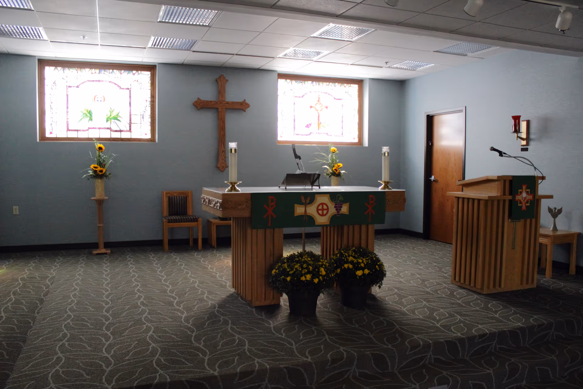 Small chapel interior with an altar, wooden cross, pulpit, stained glass windows, and floral arrangements.