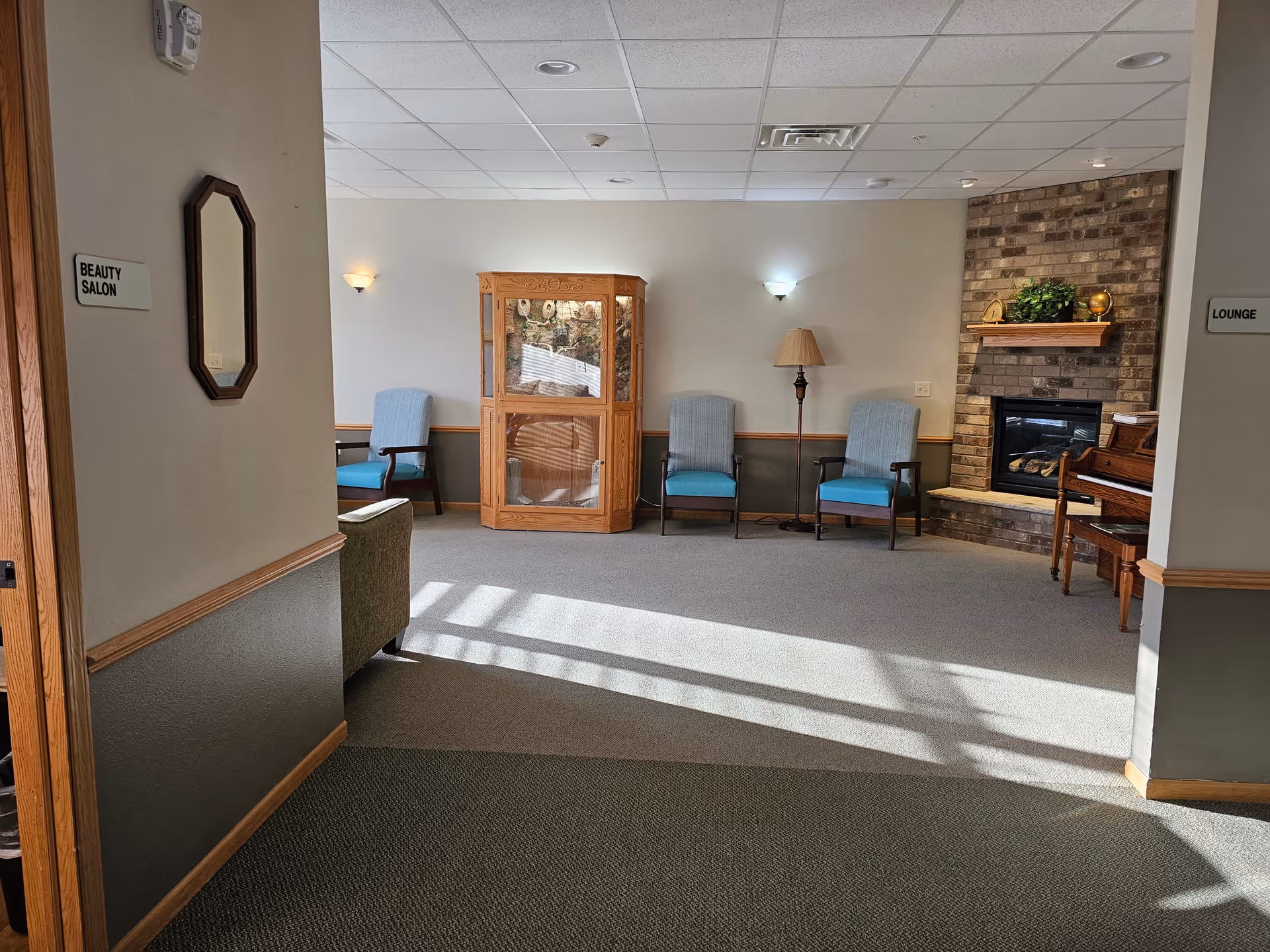 A quiet lounge area in a senior living facility with three blue cushioned chairs, a wooden display cabinet, a floor lamp, and a brick fireplace with a wooden mantel decorated with greenery and a clock. There is a piano to the right of the fireplace. The walls are painted white with a green lower half and wooden trim. Signs on the walls indicate directions to the Beauty Salon on the left and the Lounge on the right.