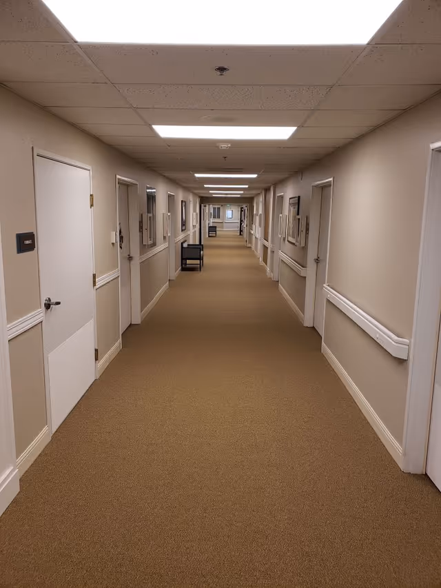 A long, well-lit hallway in a senior living facility with beige walls and carpet, white doors on both sides, handrails along the walls, and framed pictures hanging on the walls.