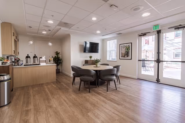 A bright and modern common area with wood flooring featuring a round table surrounded by six gray upholstered chairs. On the left side, there is a small kitchenette area with a countertop, coffee dispensers, and cabinets. The back wall has a mounted flat-screen TV above a small cabinet. Large windows and glass doors on the right side let in natural light and provide a view of an outdoor patio with tables and umbrellas.