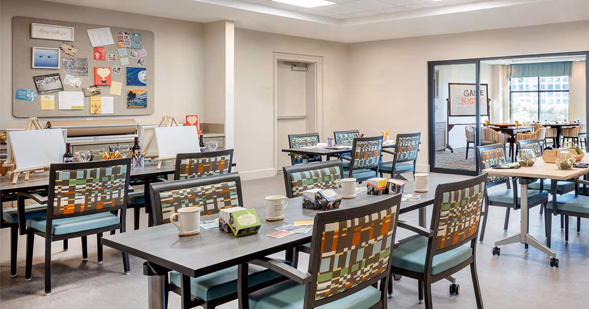 A bright, spacious activity room with multiple tables and chairs arranged for group activities. Some tables have art supplies, blank canvases on easels, and mugs. A bulletin board with various notes and pictures is mounted on the wall. Through glass doors, another room with more tables and chairs is visible, featuring a sign that reads 'Game Night'.
