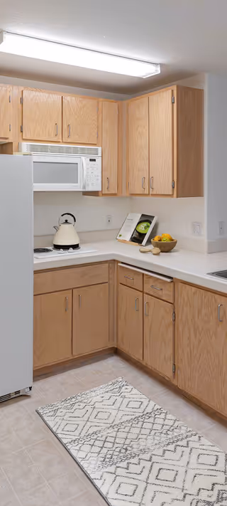 A kitchen corner with light wood cabinets, a white microwave mounted above a stove with a white kettle, a white refrigerator, a countertop with a bowl of fruit and an open cookbook, and a patterned rug on the tiled floor.