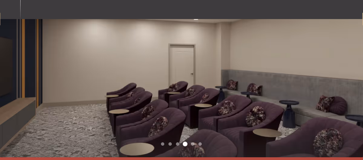 A cozy interior room with multiple purple armchairs arranged in rows, each accompanied by small round side tables. The room has a neutral color palette with beige walls, a patterned carpet, and a built-in bench along the right wall with decorative pillows. A door is visible at the back of the room.