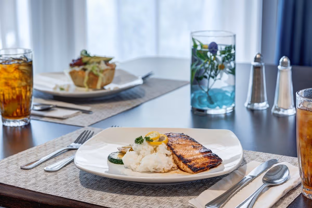 A dining table set with a plate of grilled salmon, mashed potatoes garnished with lemon slices and parsley, and a slice of zucchini. There are two glasses of iced tea, a fork, knife, and spoon on a napkin, salt and pepper shakers, and a decorative glass with water and flowers on the table. In the background, there is another plate with a salad or sandwich.