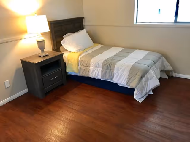 Small bedroom with a single bed, a nightstand and lamp on a hardwood floor beneath a window.