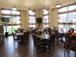 Bright dining room with several set tables, large windows with patterned curtains, pendant lights, and potted plants.