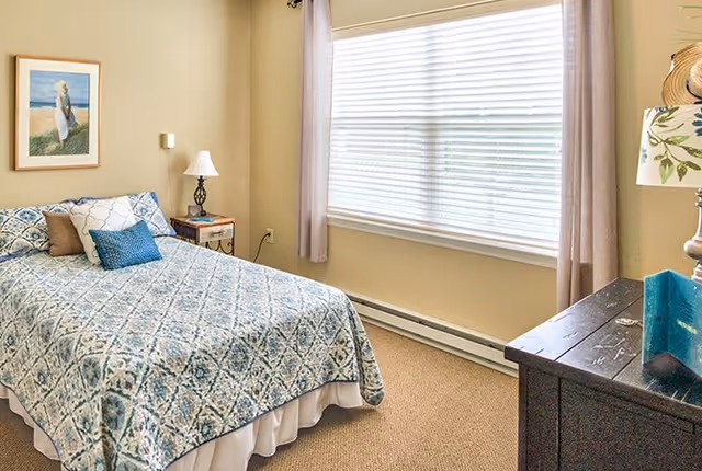 A bedroom with a large window covered by white blinds and beige curtains. The room features a bed with a blue and white patterned bedspread and several pillows. There is a nightstand with a lamp and a framed picture on the wall above the bed. A dark wooden dresser with a lamp and decorative items is positioned opposite the bed.