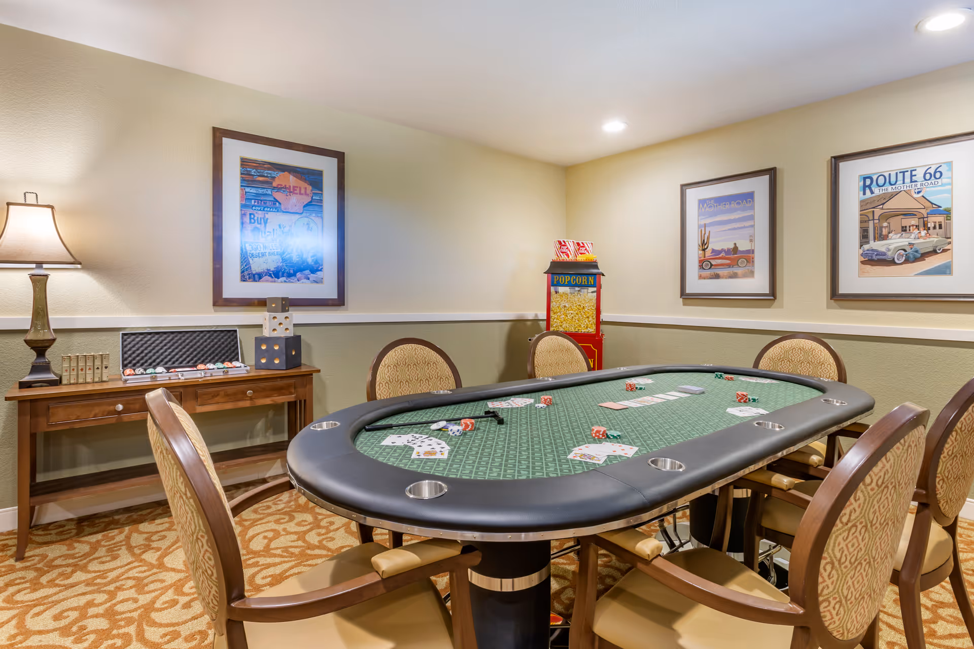 A well-lit game room with a green poker table surrounded by six cushioned chairs. Playing cards and dice are scattered on the table. Behind the table, there is a wooden console table with a lamp, poker chips, and large decorative dice. On the wall are three framed vintage-style posters, and a popcorn machine is placed in the corner.