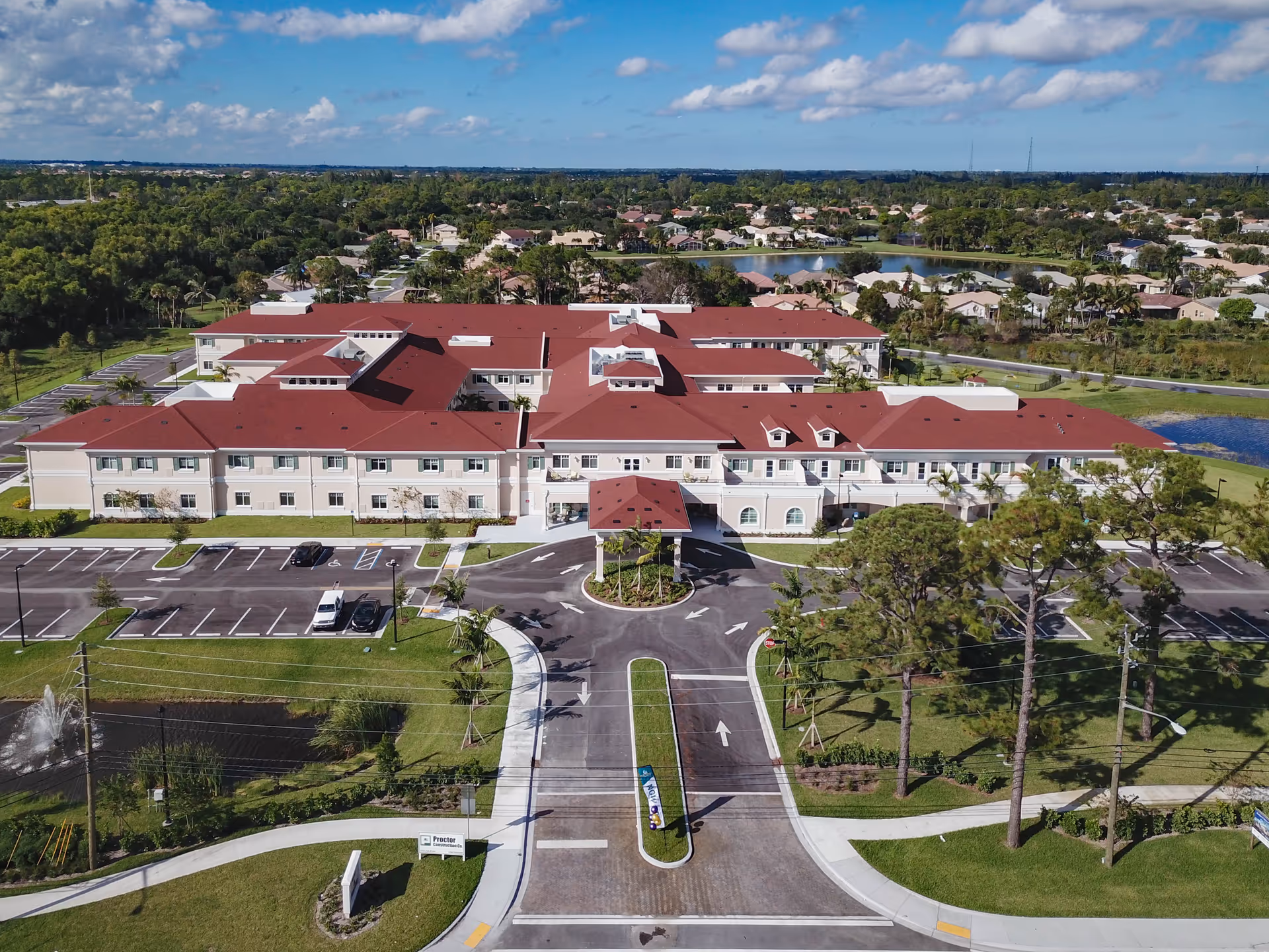 Aerial view of a large senior living building with red roofs, a circular driveway, parking lot, and surrounding trees and ponds.