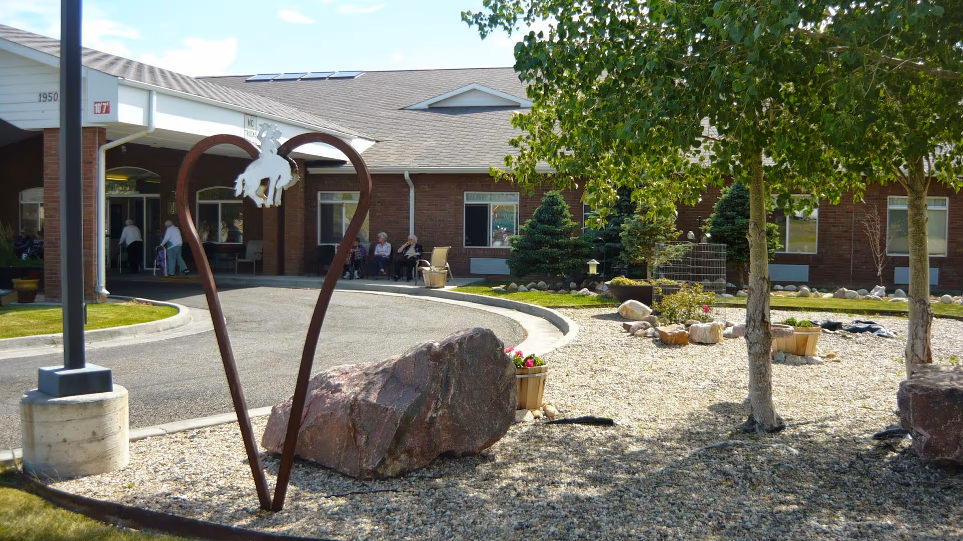 Outdoor view of Garden Square Assisted Living of Casper showing a curved driveway leading to the entrance of a brick building. There is a metal heart-shaped sculpture with a cowboy on a bucking horse in the center foreground, surrounded by rocks, small trees, and flower pots. Several elderly people are sitting near the entrance under a covered walkway.