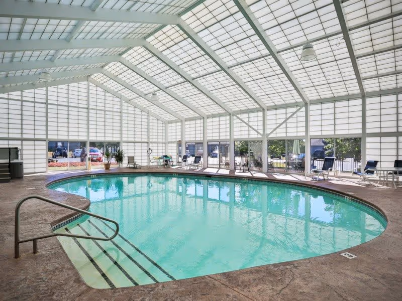 Indoor swimming pool with clear water surrounded by a textured concrete deck. The pool area is enclosed by large glass panels and a high, slanted glass ceiling allowing natural light to fill the space. Several lounge chairs and tables are arranged around the pool, with some potted plants near the glass walls.