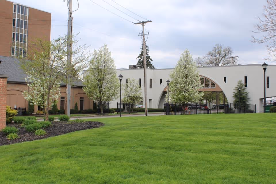 Well-maintained green lawn and landscaped trees in front of a white building with a large arched entrance and lamp posts.