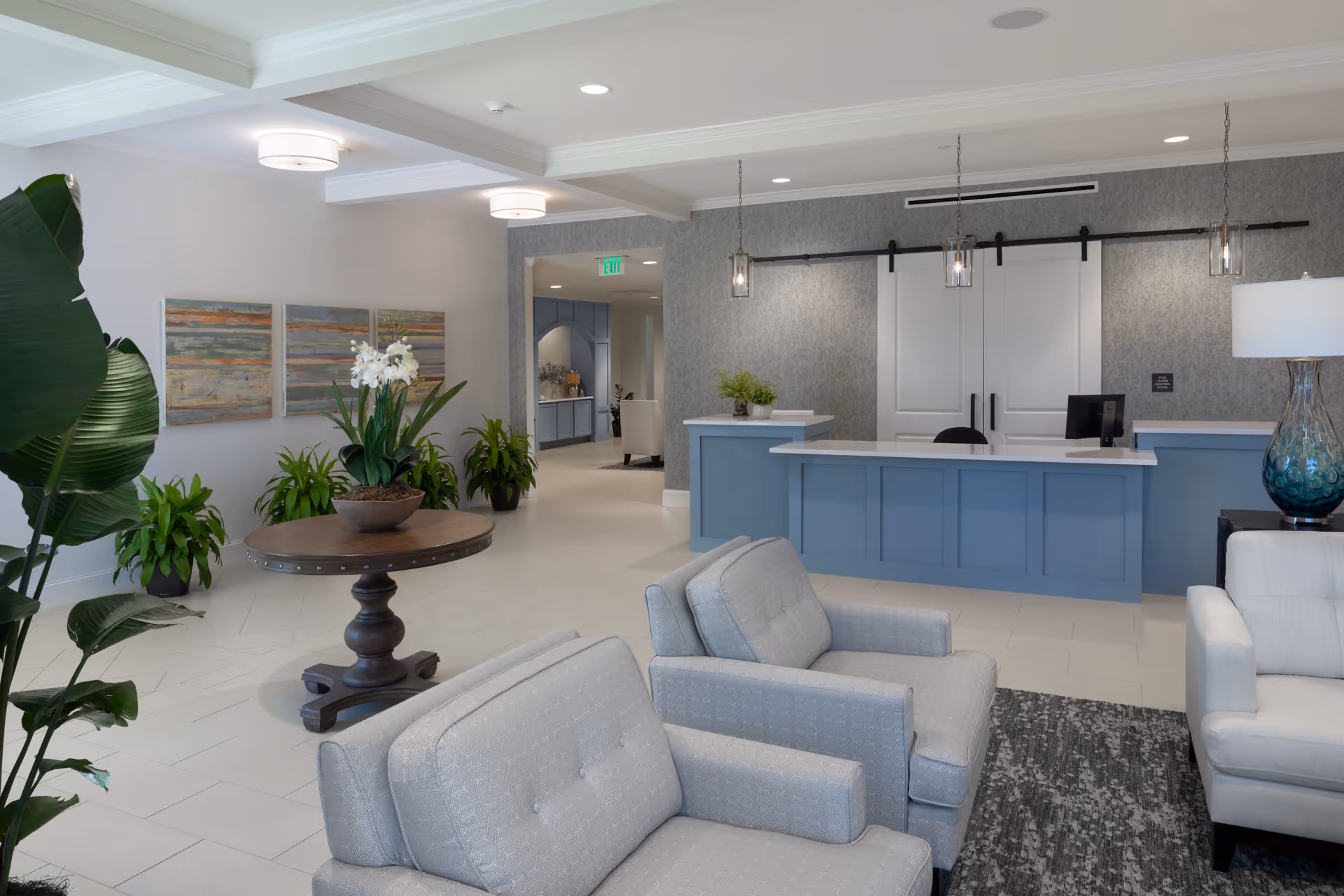 A modern and welcoming senior living facility lobby with light gray armchairs, a round wooden table with a potted orchid, several green plants, a blue reception desk with a computer, pendant lights, and abstract artwork on the wall.