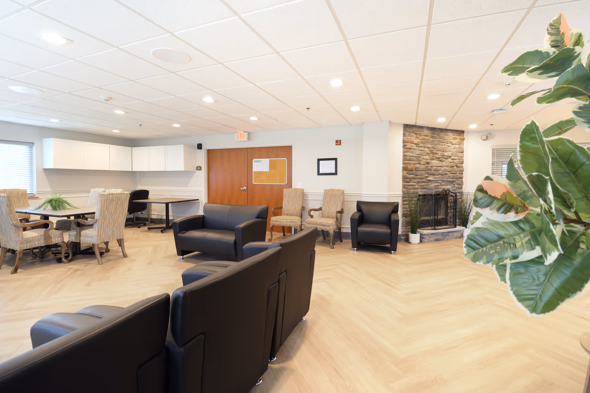 Bright communal lounge with black leather sofas, dining table and chairs, a stone fireplace, and a large potted plant.