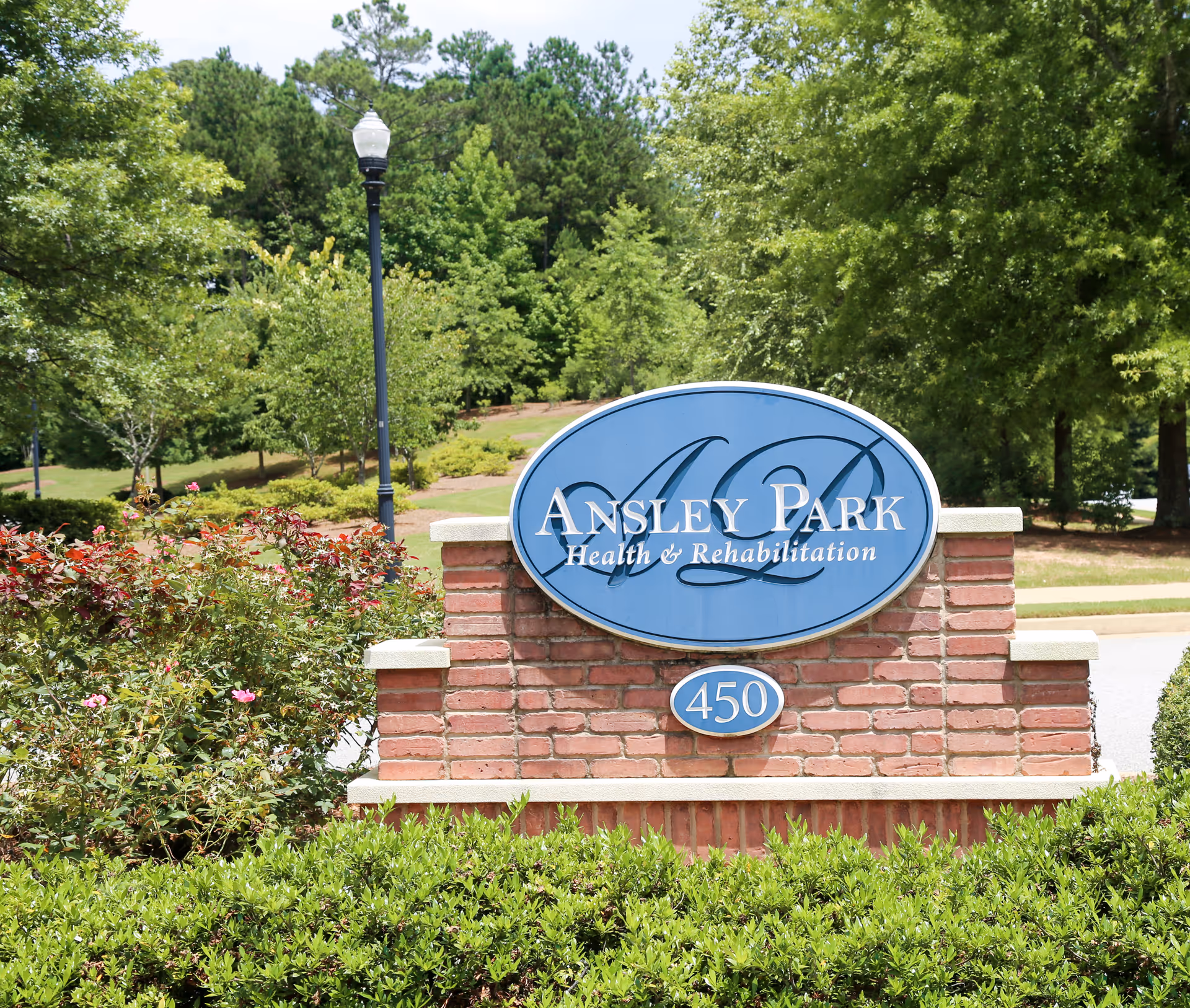 A brick sign with a blue oval plaque displaying 'Ansley Park Health & Rehabilitation' and the number 450, surrounded by green bushes and trees with a lamppost in the background.