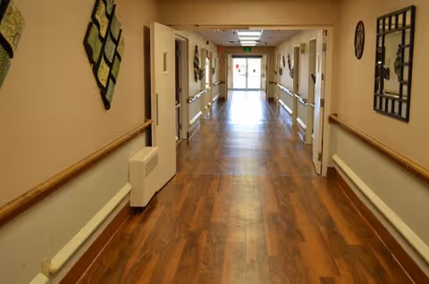 A long, well-lit hallway with wooden flooring and beige walls. The hallway has handrails on both sides and several doors along the walls. Decorative wall art and mirrors are hung on the walls. At the end of the hallway, there is a glass door letting in natural light.