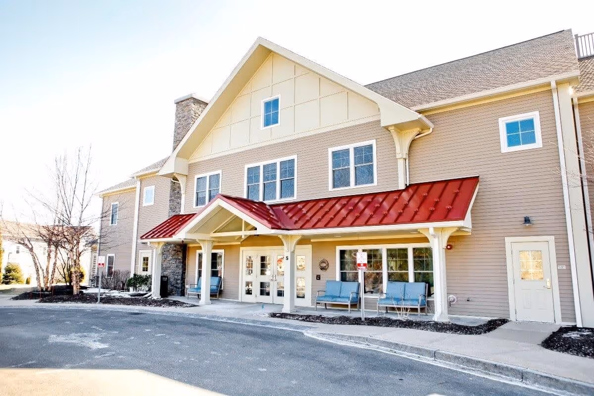Exterior front view of a senior living facility building with beige siding, a red metal awning over the entrance, white columns, and blue benches on the porch area. There are multiple windows and a stone chimney on the left side. The driveway and sidewalk lead up to the entrance.
