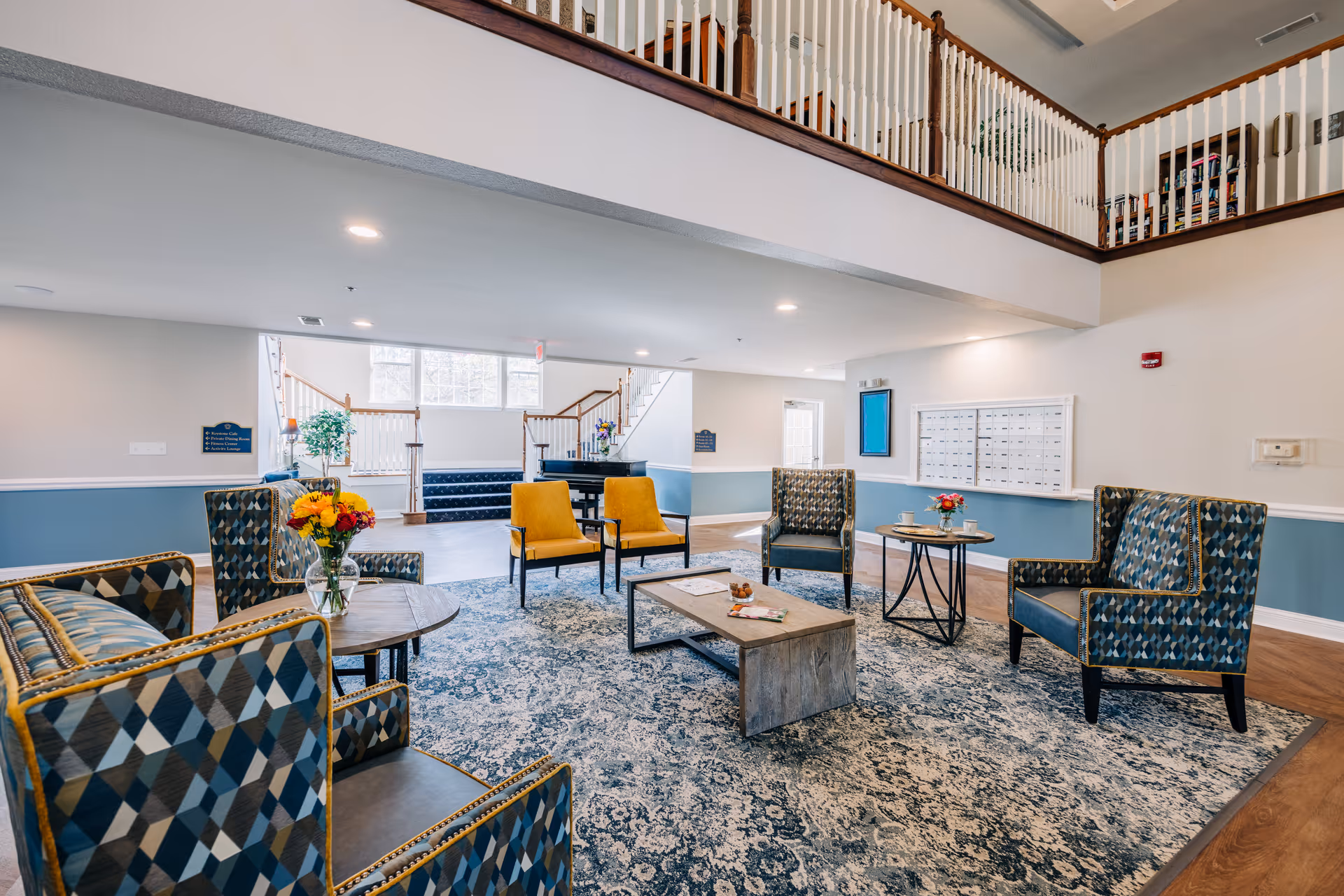 Spacious, well-lit common lounge with patterned armchairs, two yellow chairs, coffee tables, and a second-floor balcony railing.