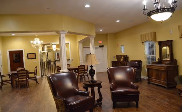 Warm, airy senior living lounge with leather chairs, tables, chandeliers, and wood flooring.