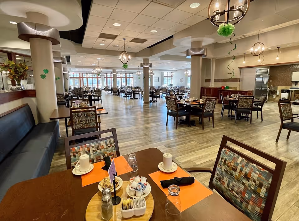 A spacious dining room in a retirement center with multiple tables and chairs arranged neatly. The tables are set with orange placemats, cups, glasses, and condiments. The room has large windows allowing natural light, decorative hanging lights, and columns with green shamrock decorations.