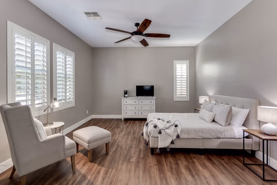 A bright and spacious bedroom with light gray walls and wood flooring. The room features a large bed with white bedding and decorative pillows, two bedside tables with lamps, a white dresser with a small TV and coffee maker on top, a comfortable armchair with an ottoman, and three windows with white plantation shutters. A ceiling fan with wooden blades is mounted on the ceiling.