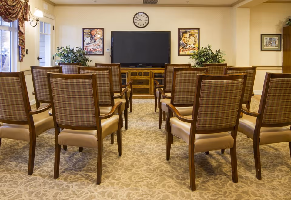 A small theater or media room with rows of wooden chairs featuring plaid upholstery, facing a large flat-screen TV mounted on a wooden cabinet. The room has beige patterned carpet, light-colored walls, two framed vintage movie posters on either side of the TV, a clock above the TV, and potted plants on either side of the cabinet.