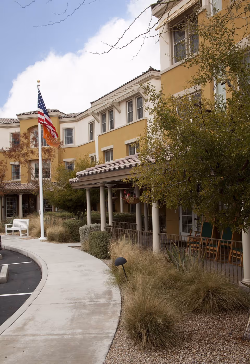 Front exterior of a multi-story yellow senior living building with a covered porch, landscaping, and an American flag.