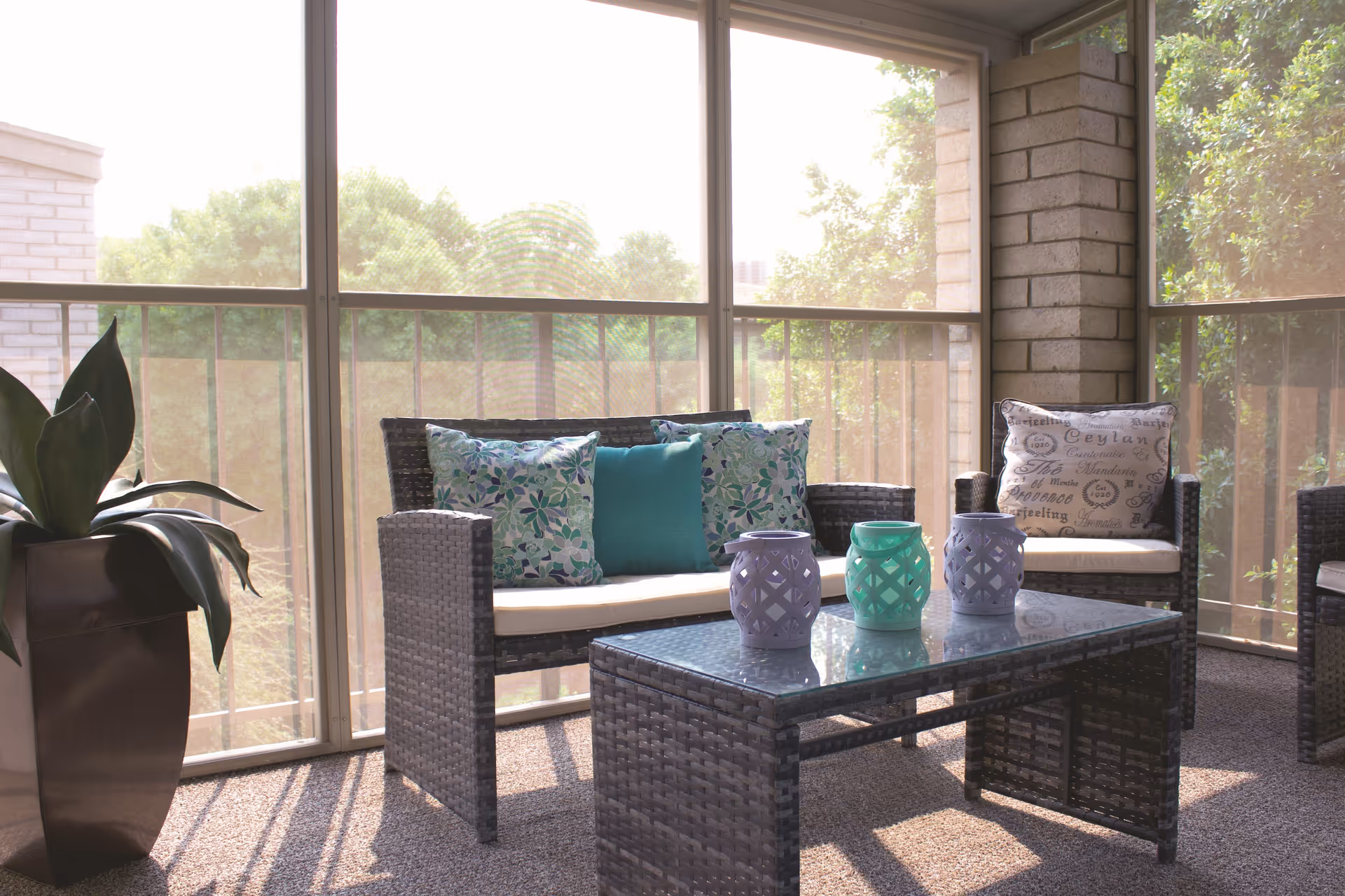 A sunlit indoor seating area with wicker furniture including a loveseat and chairs adorned with decorative cushions. A glass-top coffee table holds three decorative lanterns in pastel colors. Large windows with a view of green trees surround the space, and a large potted plant is placed to the left.