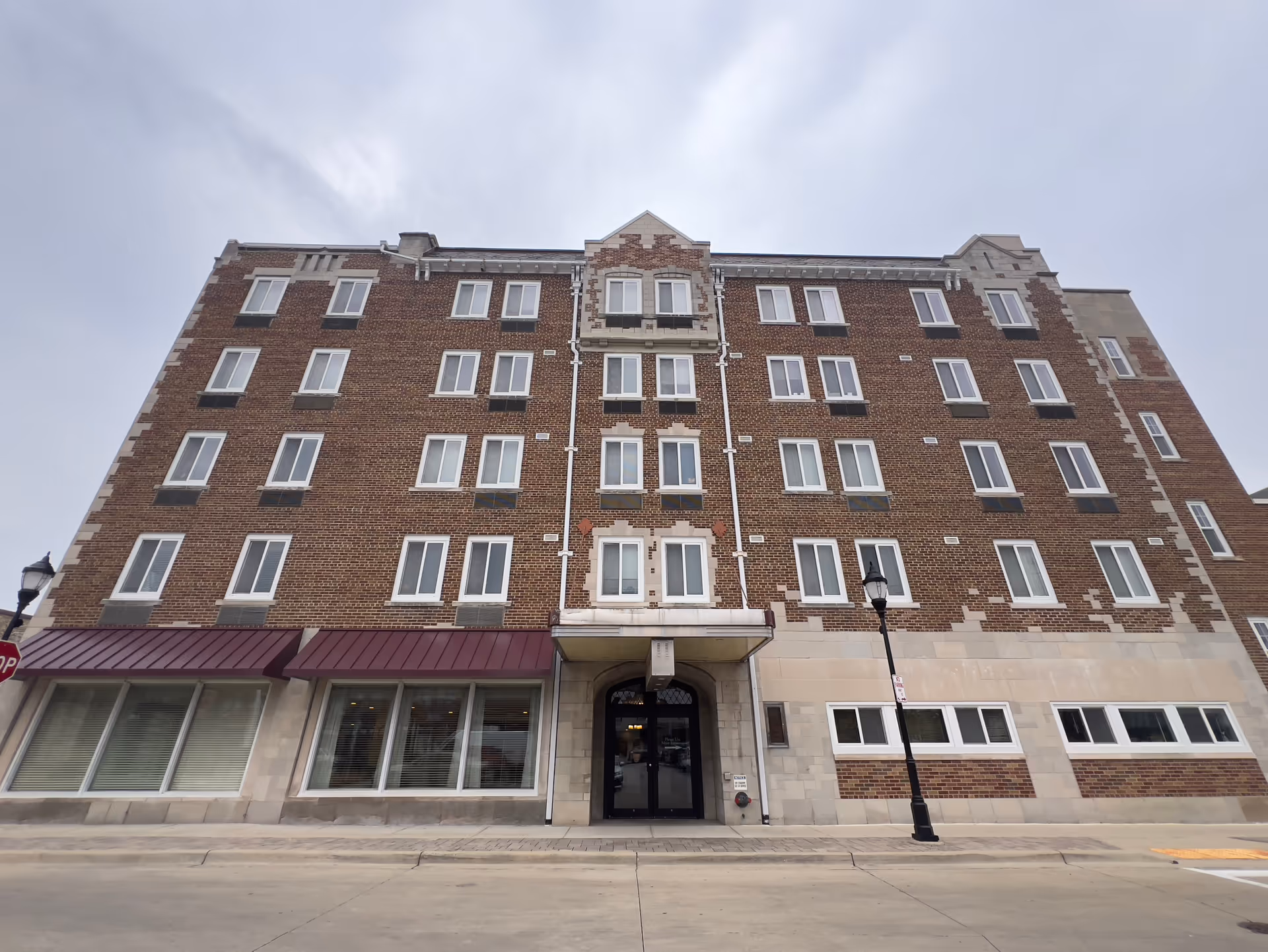 Front exterior view of a multi-story brick building with numerous windows and a central entrance under a small awning. The building has decorative brickwork and stone accents, with a street lamp and sidewalk in front.