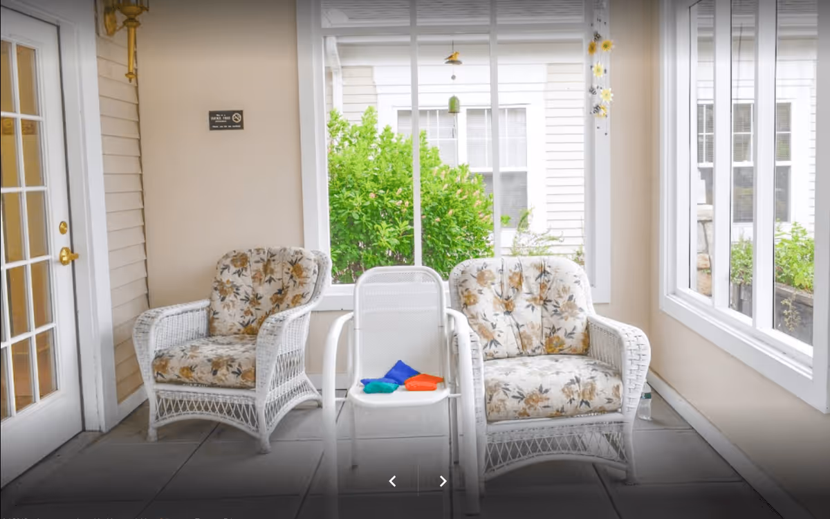 A bright indoor seating area with two white wicker armchairs featuring floral cushions and a white metal chair between them. The area is surrounded by large windows showing green bushes outside. There is a glass door to the left and a beige wall with a small sign.