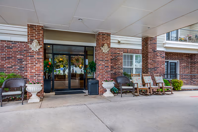 Covered brick entrance with glass double doors, wreaths, potted plants, and seating under a canopy.