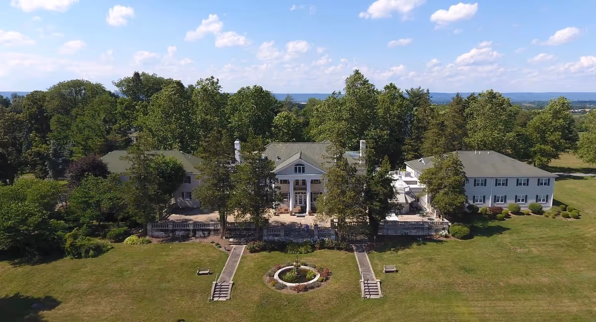 A large estate building with classical architecture featuring tall white columns at the entrance, surrounded by trees and greenery under a blue sky with scattered clouds. There is a circular garden feature with a fountain in front of the building, and two pathways leading up to the entrance with benches on either side.