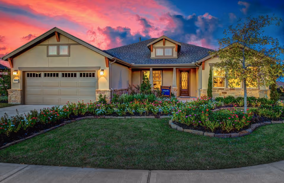 A single-story house with a two-car garage, a well-maintained front yard with flower beds and a small tree, and a vibrant sunset sky in the background.