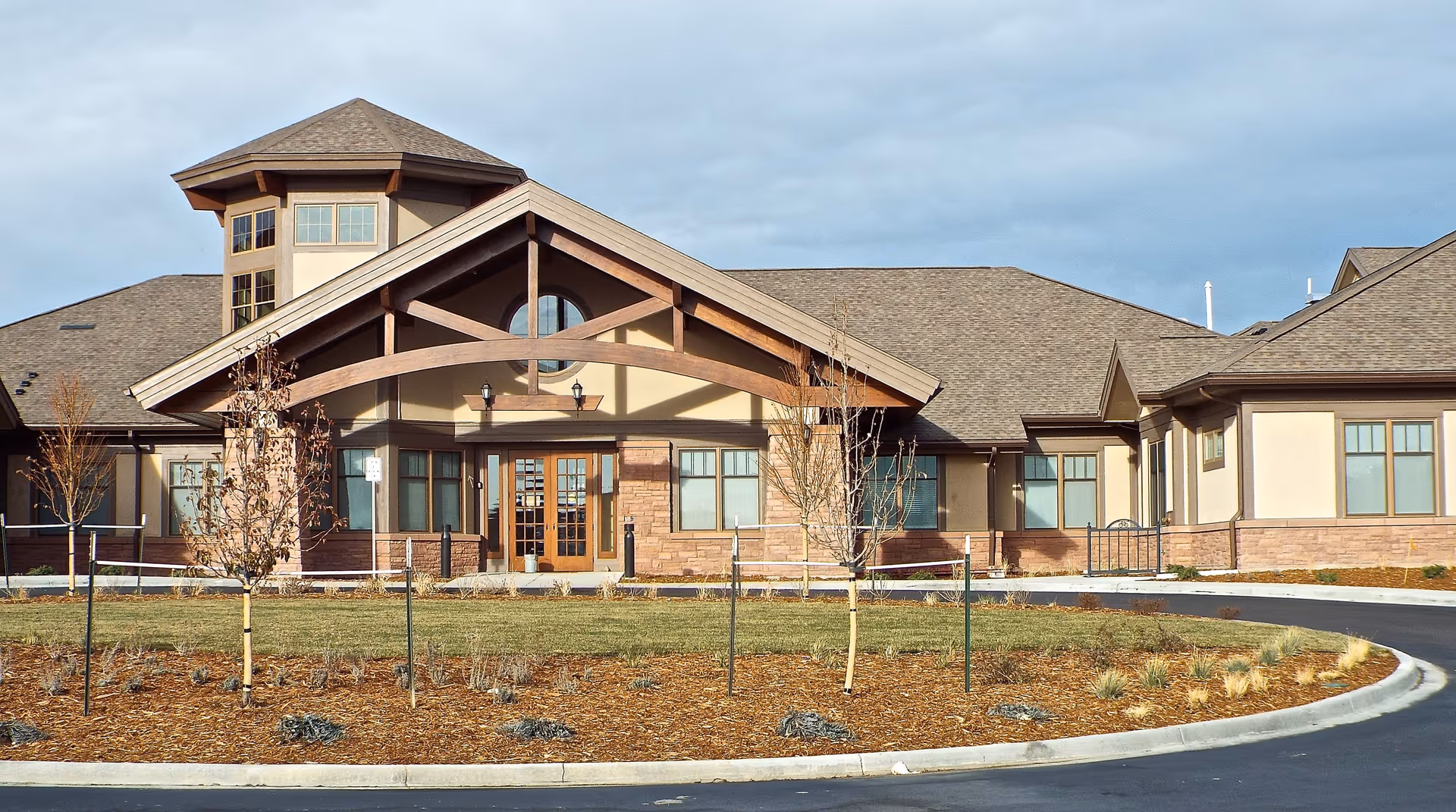 Front exterior view of a single-story building with a peaked roof and large wooden entrance structure, surrounded by a landscaped area with young trees and mulch.