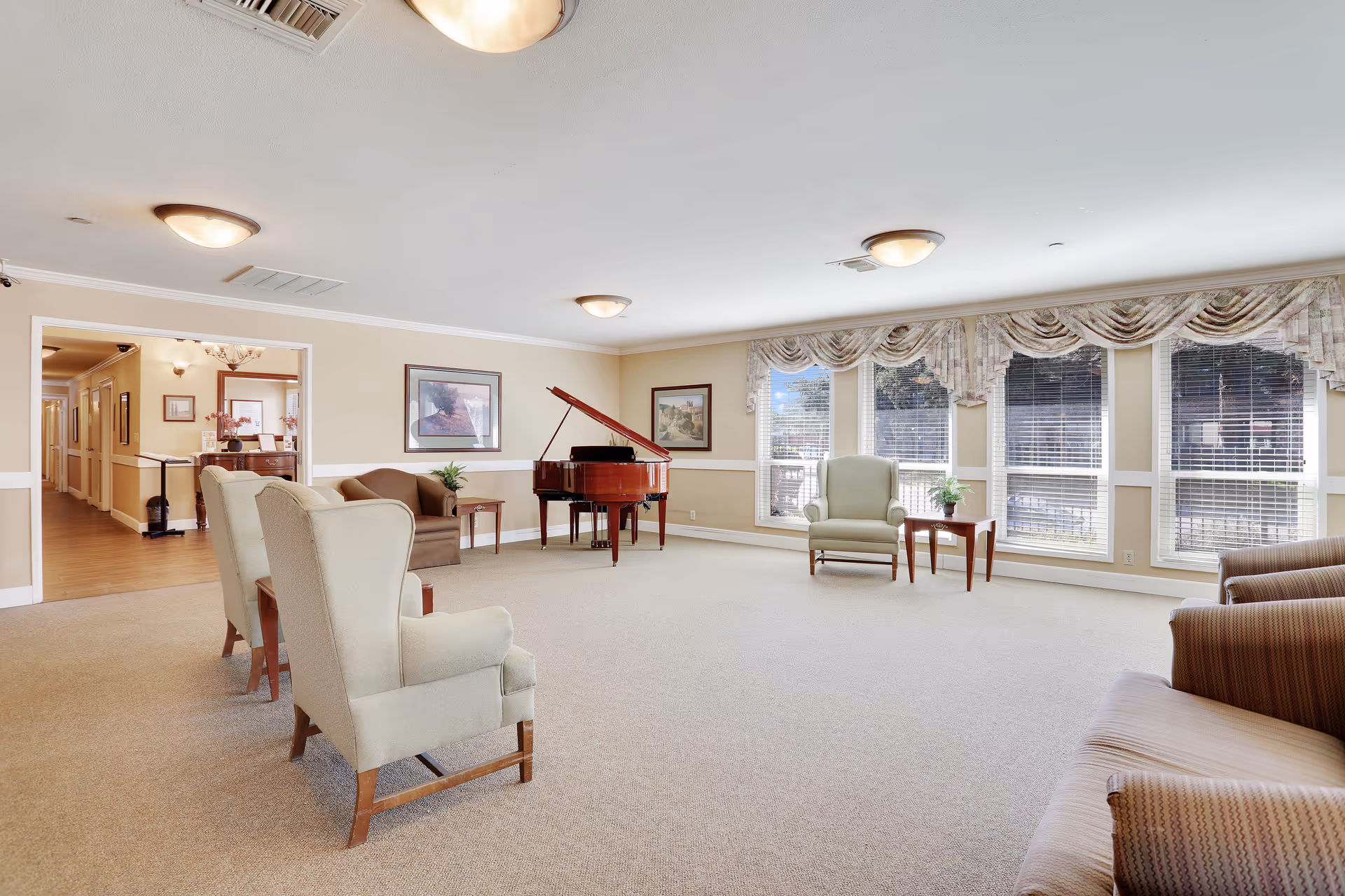 A spacious and well-lit assisted living facility common room with beige walls and carpet. The room features several upholstered chairs arranged in a semi-circle, a brown sofa, a grand piano, and large windows with floral valances letting in natural light. There are framed pictures on the walls and small tables with plants.