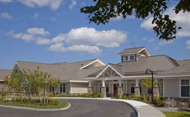 Exterior view of a senior living facility building with a covered entrance, stone pillars, and a curved driveway. The sky is blue with some clouds, and there are trees and landscaping around the building.