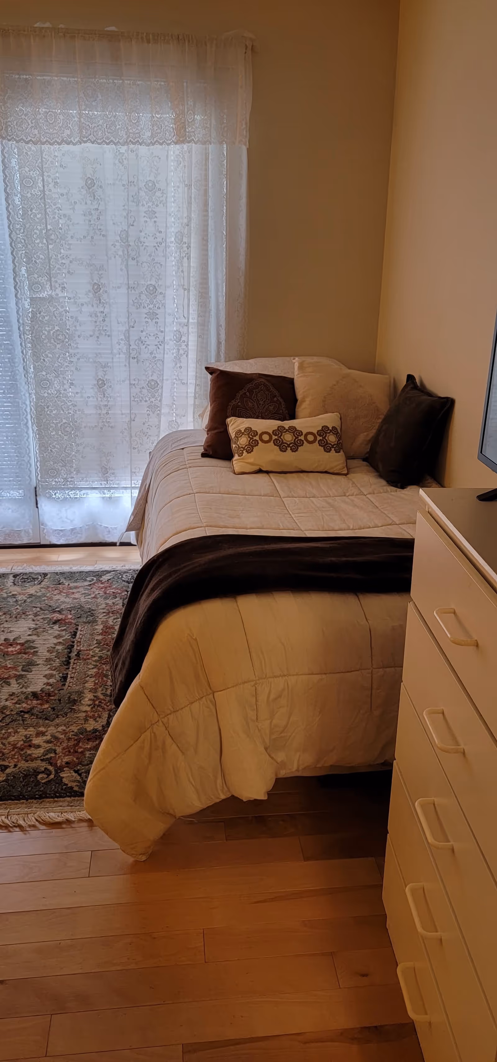 A small bedroom with a single bed dressed with pillows and a throw, a dresser, and a lace-curtained window.