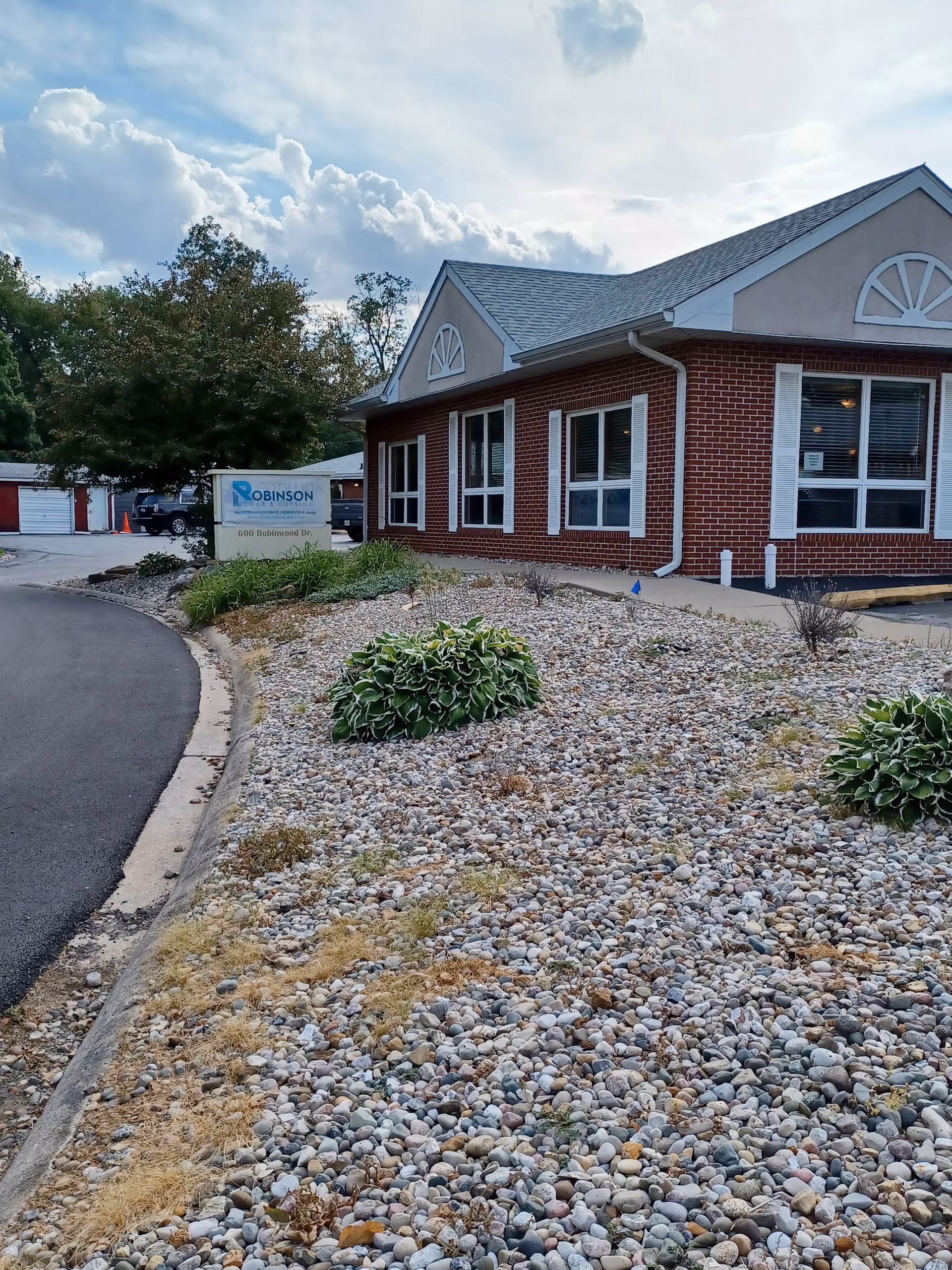 Exterior view of a brick building with white window shutters and a gray roof, surrounded by a landscaped area with rocks and some green bushes. A sign near the building reads 'Robinson Rehab and Nursing LLC' with the address '600 Robinwood Dr.'. The sky is partly cloudy.